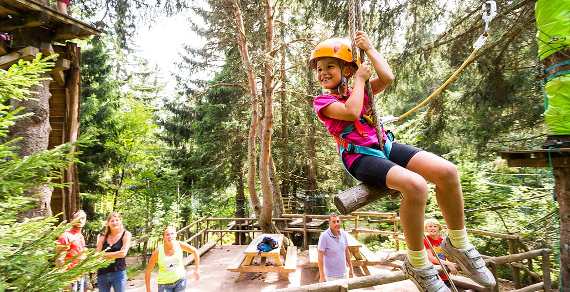 Une jeune fille souriante, portant un casque de sécurité, fait de la tyrolienne dans une forêt, observée par un groupe de personnes en bas, parmi les arbres et des plateformes en bois.