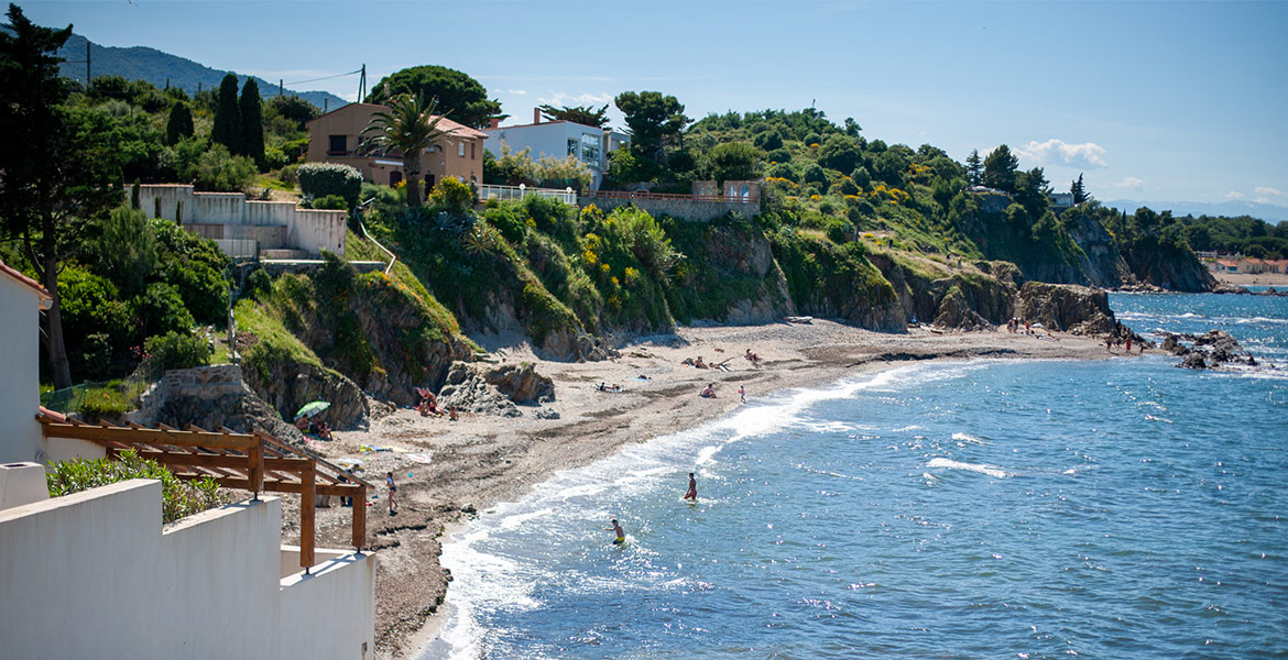 Des personnes profitent d'une plage bordée de maisons surplombant une colline verdoyante. La mer bleue et le sable clair créent une atmosphère estivale et relaxante.