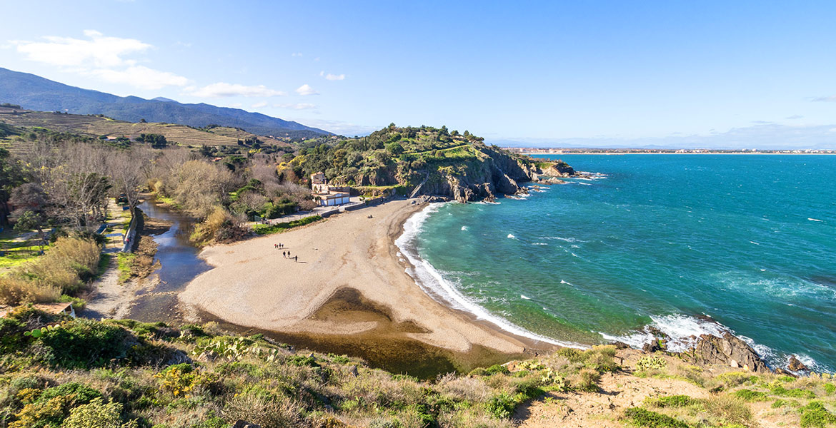 Une plage isolée en forme de croissant est entourée de collines verdoyantes et d'une mer turquoise. Quelques personnes se promènent sur le sable sous un ciel bleu lumineux.