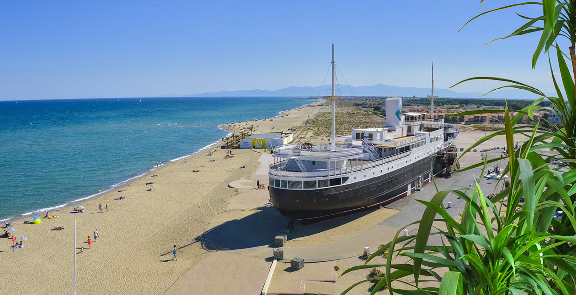 Un grand bateau est échoué sur une plage de sable fin, où des personnes profitent du soleil et de la mer. En arrière-plan, on peut voir des montagnes et un ciel bleu clair.