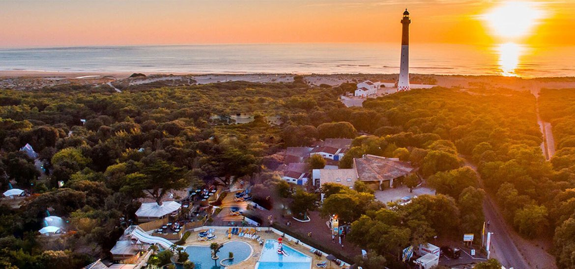 Vue aérienne d'un phare en bord de mer au coucher du soleil, avec une forêt dense et des installations de loisirs en premier plan.