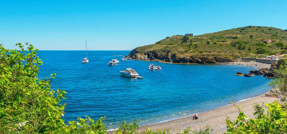 Des bateaux de plaisance sont ancrés dans une baie aux eaux bleues cristallines, bordée d'une plage de galets et de collines verdoyantes sous un ciel dégagé dans les Pyrénées Orientales.