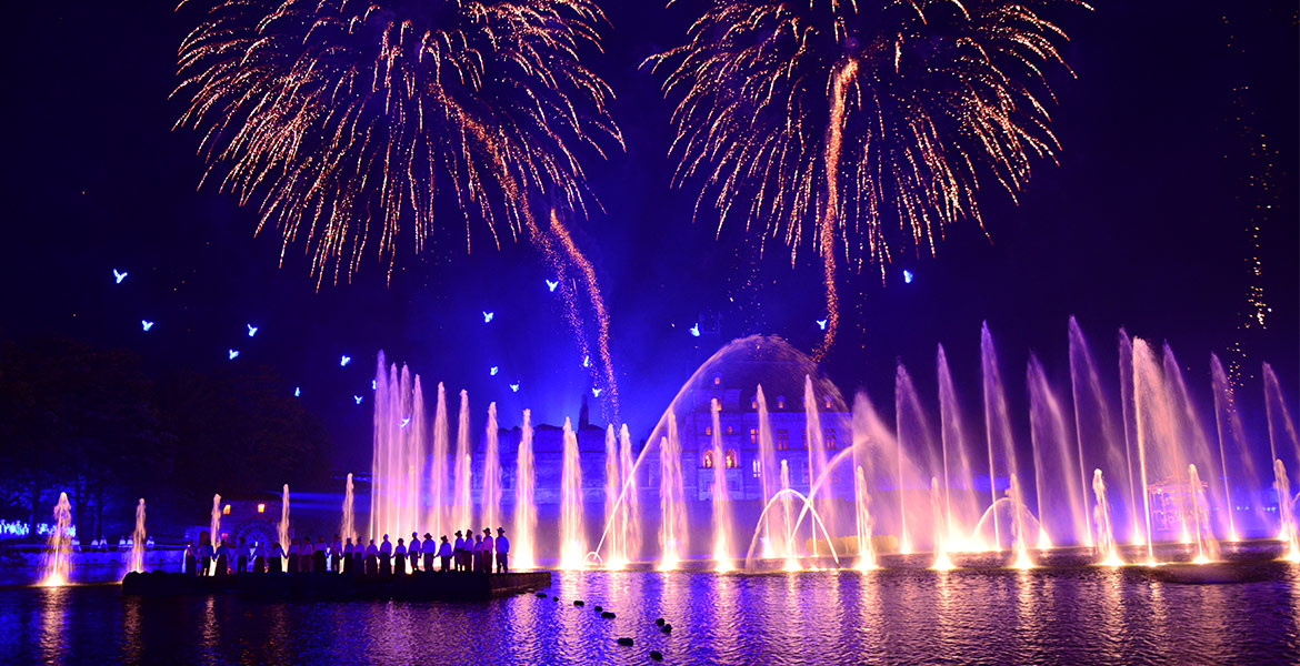 Le spectacle de la Cinéscénie au Puy du Fou en Vendée avec des feux d'artifice éclatant dans le ciel et des fontaines lumineuses jaillissant de l'eau, illuminant une scène devant une foule de spectateurs.