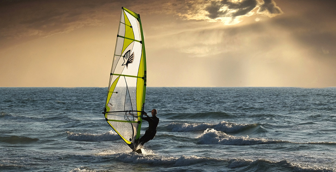 Un homme fait de la planche à voile sur l'océan, sous un ciel nuageux avec le soleil couchant en arrière-plan.