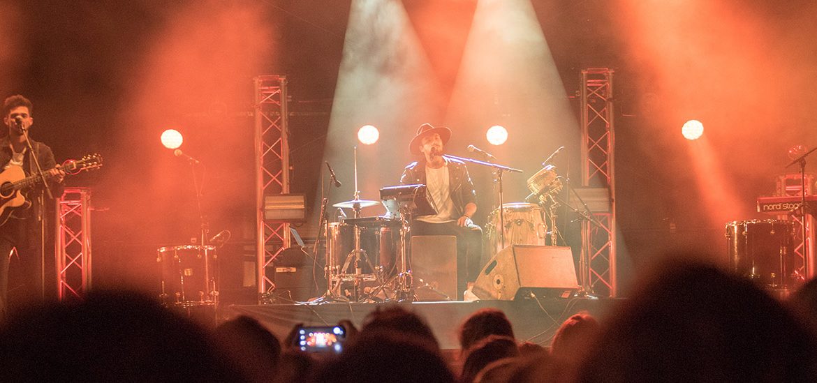 Groupe de musique se produisant sur scène avec des éclairages orangés et une foule de spectateurs au premier plan. Un musicien joue de la guitare à gauche, un batteur au centre, et un autre musicien au clavier à droite.