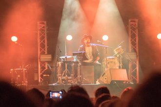 Groupe de musique se produisant sur scène avec des éclairages orangés et une foule de spectateurs au premier plan. Un musicien joue de la guitare à gauche, un batteur au centre, et un autre musicien au clavier à droite.