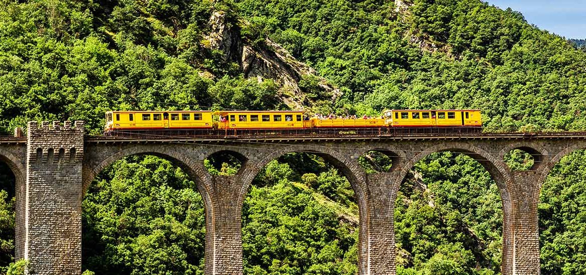 Train jaune traversant un viaduc en pierre au milieu de montagnes verdoyantes dans les Pyrénées Orientales.