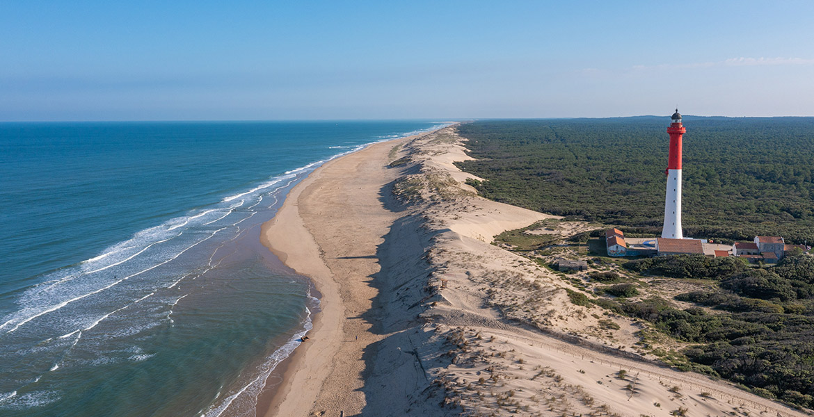 Vue aérienne du Phare e la Coubre situé à la Palmyre sur une dune de sable, avec une longue plage de sable fin s'étendant à perte de vue le long de l'océan. À gauche, l'eau bleue de l'océan Atlantique et à droite, une vaste forêt verdoyante.