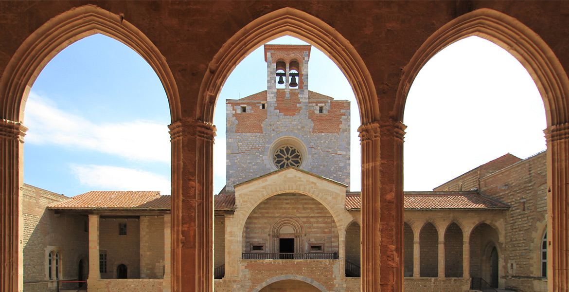 Vue à travers des arches du cloître d'une abbaye, montrant une façade en pierre avec une grande rosace et un clocher à deux cloches, sous un ciel bleu.