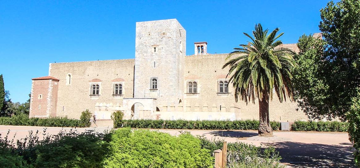 Un bâtiment historique en pierre avec des fenêtres anciennes et une grande cour, entouré de végétation et d'arbres, sous un ciel bleu clair.