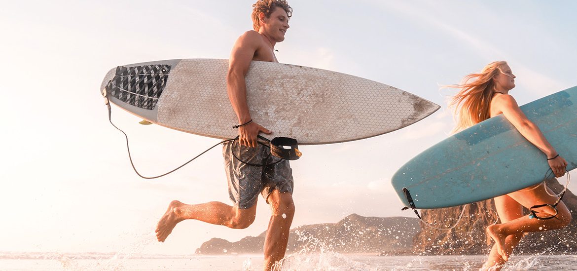 Un homme et une femme courent dans l'eau en tenant des planches de surf, se dirigeant vers l'océan sous un ciel clair.