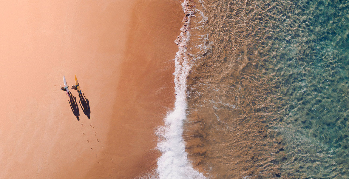 Vue aérienne de deux surfeurs marchant sur une plage de sable, portant leurs planches de surf et laissant des empreintes derrière eux, avec l'eau claire de la mer s'écrasant doucement sur le rivage à droite de l'image.