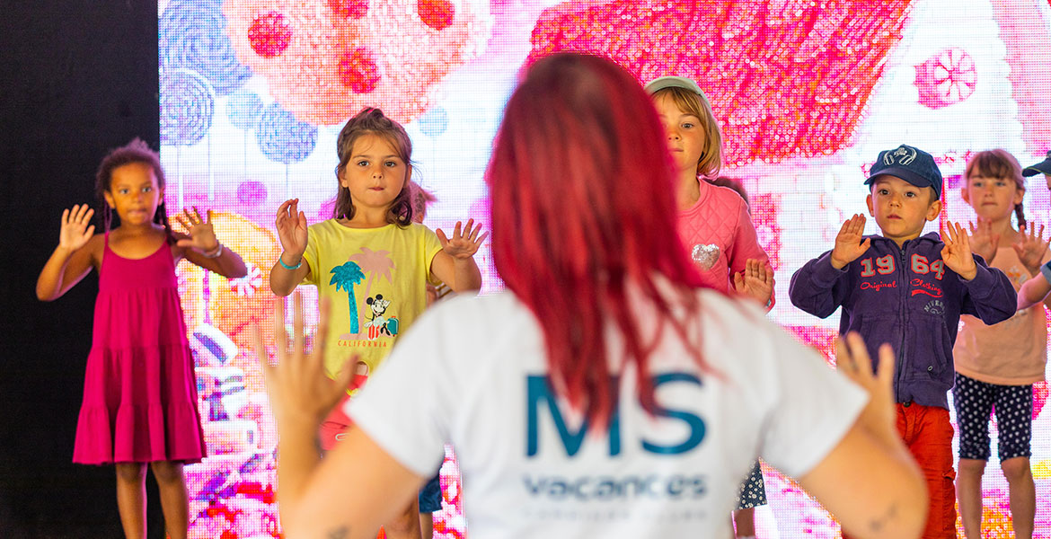 Une animatrice avec un t-shirt "MS Vacances" guide un groupe d'enfants dans une activité sur scène, avec un décor coloré en arrière-plan.