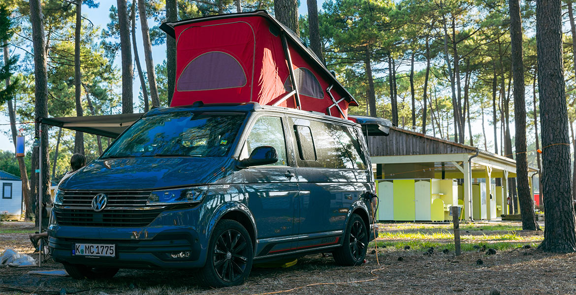 Un van Volkswagen bleu avec un toit relevé rouge, garé dans un camping forestier près de bâtiments de services.