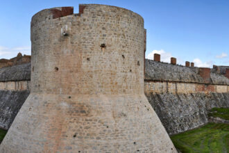 Vue panoramique de la forteresse de Sales avec une tour massive et des remparts, entouré de douves verdoyantes sous un ciel dégagé.