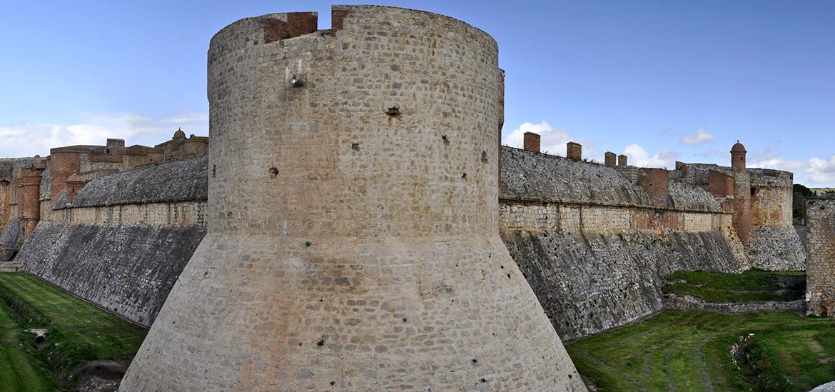 Vue panoramique de la forteresse de Sales avec une tour massive et des remparts, entouré de douves verdoyantes sous un ciel dégagé.