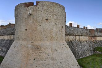 Vue panoramique de la forteresse de Sales avec une tour massive et des remparts, entouré de douves verdoyantes sous un ciel dégagé.