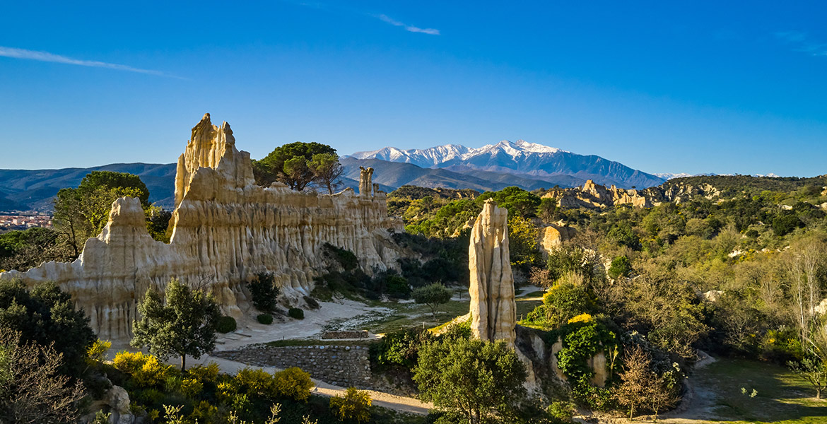 Paysage de formations rocheuses spectaculaires des Orgues d'Ille-sur-Têt avec en arrière-plan des montagnes enneigées sous un ciel bleu.