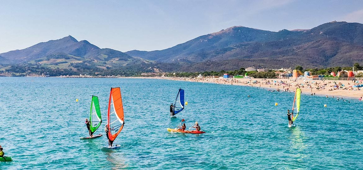 Plage d'Argeles-sur-mer animée avec des gens pratiquant le kayak et la planche à voile dans une eau turquoise, des montagnes en arrière-plan sous un ciel bleu clair.