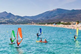 Plage d'Argeles-sur-mer animée avec des gens pratiquant le kayak et la planche à voile dans une eau turquoise, des montagnes en arrière-plan sous un ciel bleu clair.