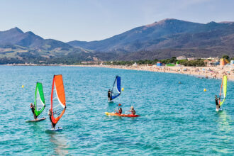 Plage d'Argeles-sur-mer animée avec des gens pratiquant le kayak et la planche à voile dans une eau turquoise, des montagnes en arrière-plan sous un ciel bleu clair.