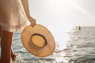 Une personne en robe blanche tenant un chapeau de paille à la main, marchant près de l'eau au coucher du soleil.