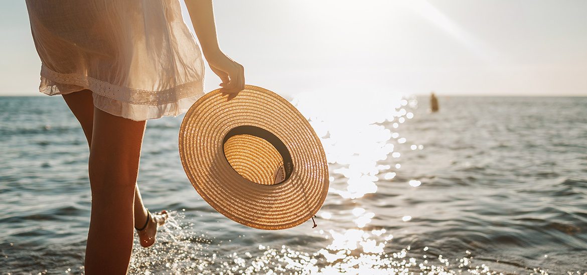 Une personne en robe blanche tenant un chapeau de paille à la main, marchant près de l'eau au coucher du soleil.