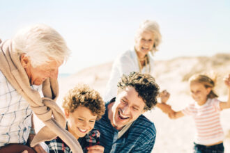 Une famille s'amusant sur une plage ensoleillée. Un grand-père tient un ballon tandis que ses petits-enfants essaient de l'attraper, sous le regard souriant des parents.