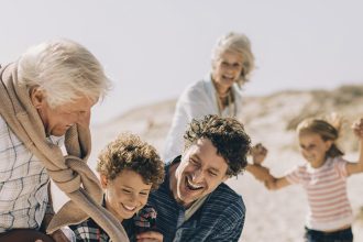 Une famille s'amusant sur une plage ensoleillée. Un grand-père tient un ballon tandis que ses petits-enfants essaient de l'attraper, sous le regard souriant des parents.