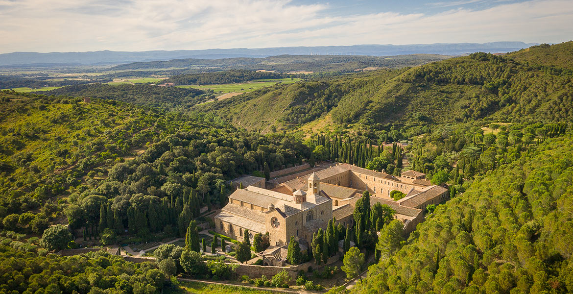 Vue aérienne de l'Abbaye de Fontfroide entourée de collines verdoyantes et de forêts. L'architecture historique de l'abbaye se détache au milieu de la nature luxuriante.