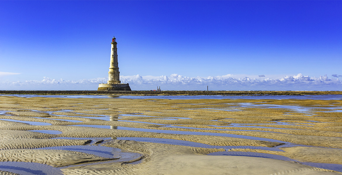 Phare de Cordouan avec des motifs de marée basse, entouré par l'océan sous un ciel bleu clair avec des nuages à l'horizon.