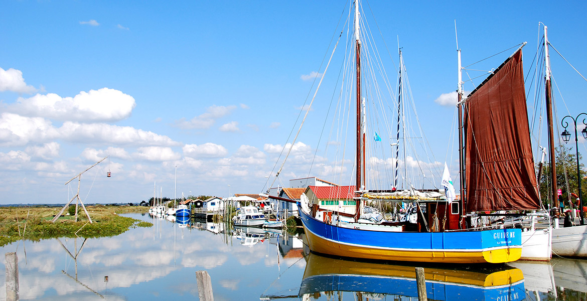 Bateau de pêche coloré amarré dans le port de Mornac sur Seudre, avec des maisons et d'autres bateaux en arrière-plan sous un ciel bleu parsemé de quelques nuages blancs.