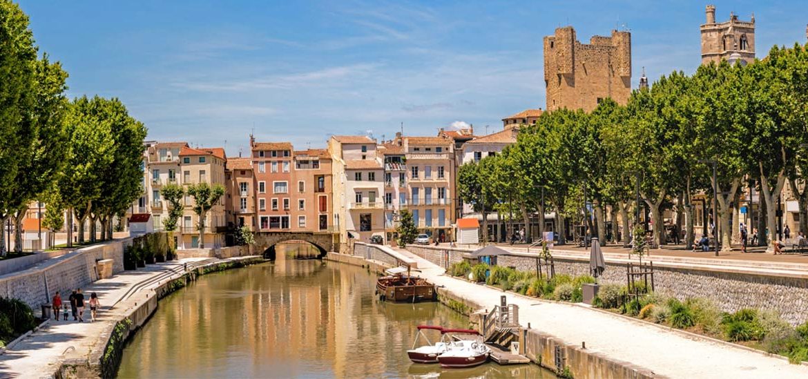 Canal bordé de bâtiments colorés et d'arbres à Narbonne dans l'Aude avec des bateaux amarrés, des passants se promenant sur les quais, et un ciel bleu clair.