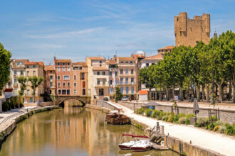 Canal bordé de bâtiments colorés et d'arbres à Narbonne dans l'Aude avec des bateaux amarrés, des passants se promenant sur les quais, et un ciel bleu clair.