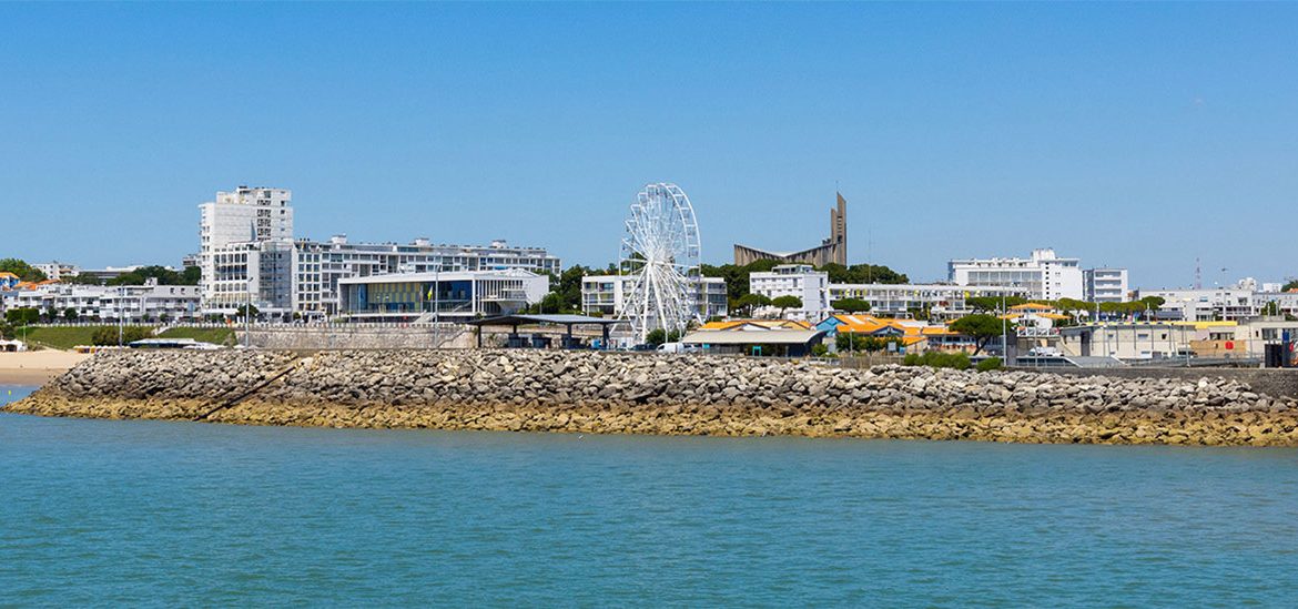 Vue de la ville de Royan depuis l'eau, avec des bâtiments modernes, une grande roue et une jetée en pierre sous un ciel bleu clair.