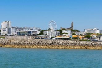 Vue de la ville de Royan depuis l'eau, avec des bâtiments modernes, une grande roue et une jetée en pierre sous un ciel bleu clair.