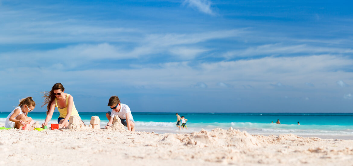 Famille jouant dans le sable sur une plage ensoleillée avec des gens se baignant dans l'eau turquoise en arrière-plan sous un ciel bleu avec quelques nuages.