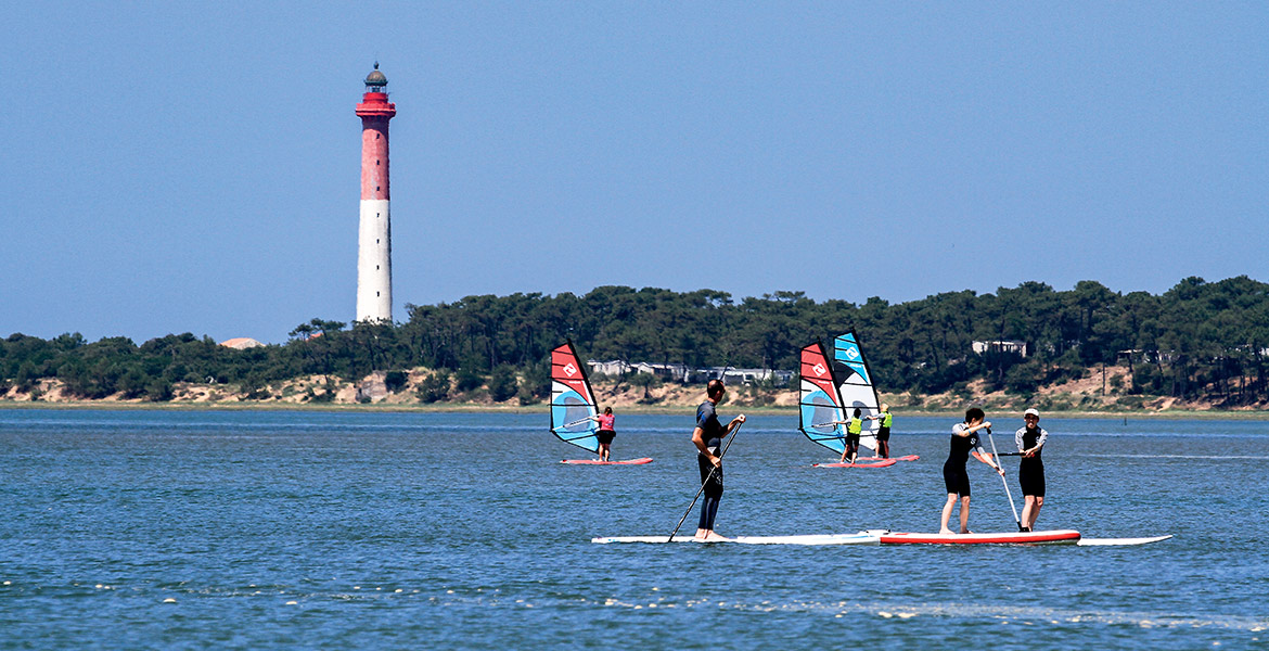 Personnes pratiquant le paddle et la planche à voile sur l'eau avec le Phare de la Coubre en arrière-plan, entouré de végétation dense sous un ciel bleu clair.