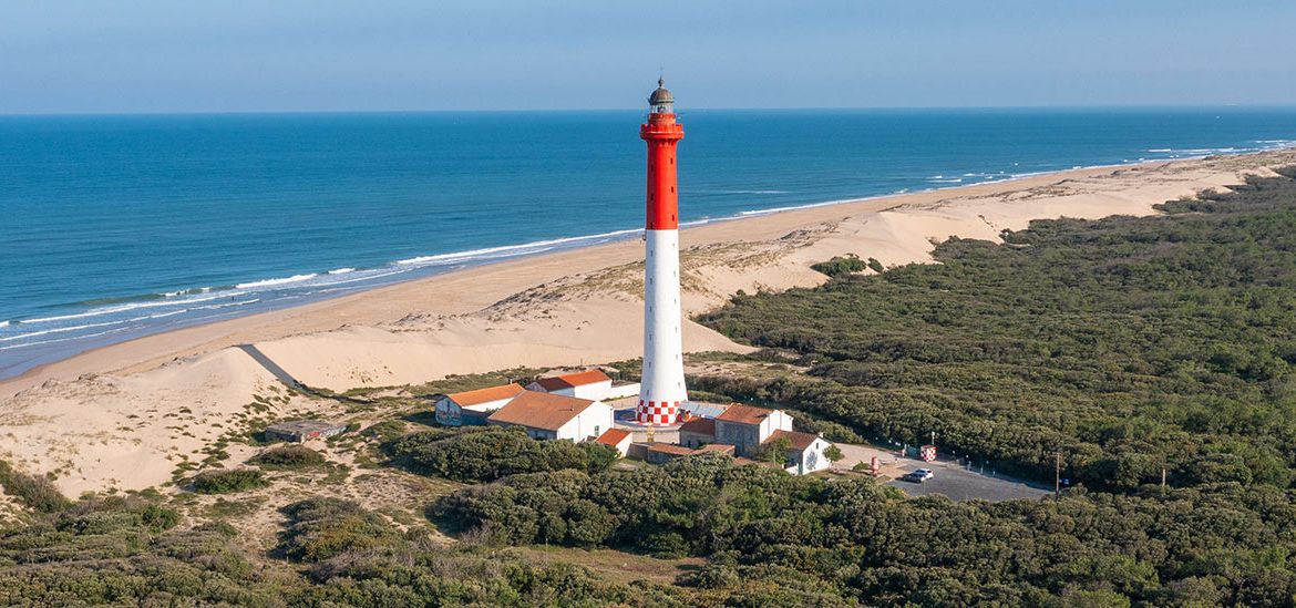 Vue aérienne du phare de la Côte Sauvage, rouge et blanc, situé près de dunes de sable et entouré de végétation dense avec l'océan Atlantique en arrière-plan sous un ciel clair.