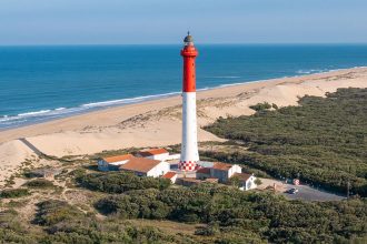 Vue aérienne du phare de la Côte Sauvage, rouge et blanc, situé près de dunes de sable et entouré de végétation dense avec l'océan Atlantique en arrière-plan sous un ciel clair.