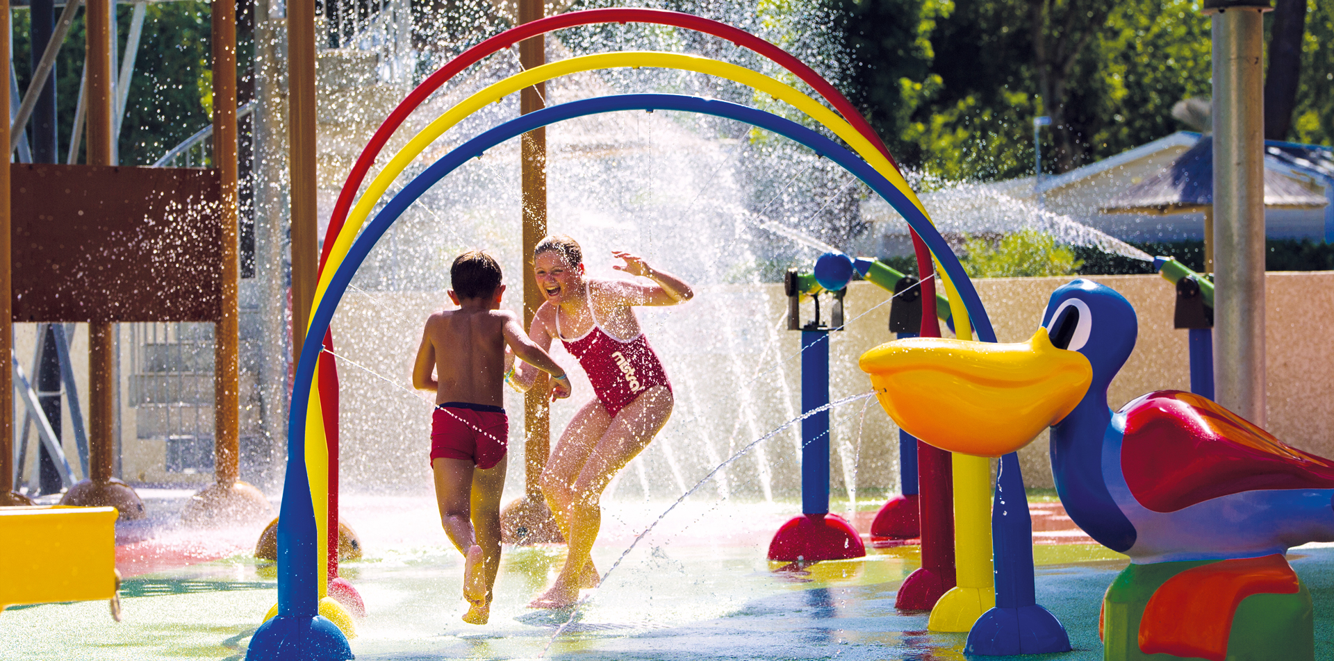 Enfants jouant et courant à travers des jets d'eau colorés dans l'aire de jeux aquatique du Camping Club MS Le Littoral sous un soleil éclatant.