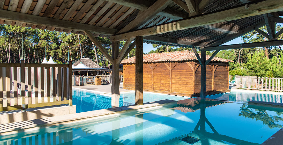 Piscine du Camping Campéole Les Tourterelles 4 étoiles extérieure partiellement couverte par une structure en bois, entourée de cabanes en bois et de végétation dense sous un ciel bleu.