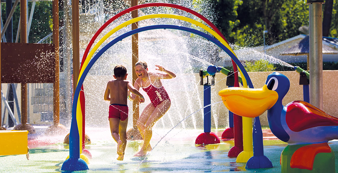 Enfants jouant et courant à travers des jets d'eau colorés dans l'aire de jeux aquatique du Camping Club MS Le Littoral sous un soleil éclatant.