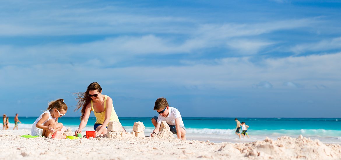 Famille jouant dans le sable sur une plage ensoleillée avec des gens se baignant dans l'eau turquoise en arrière-plan sous un ciel bleu avec quelques nuages.