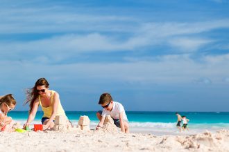 Famille jouant dans le sable sur une plage ensoleillée avec des gens se baignant dans l'eau turquoise en arrière-plan sous un ciel bleu avec quelques nuages.