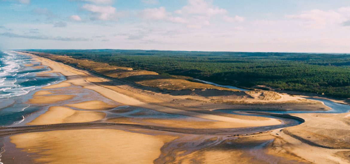 Vue aérienne de la plage de Saint Girons étendue avec des dunes de sable, un cours d'eau serpentant et une forêt verdoyante en arrière-plan sous un ciel partiellement nuageux.