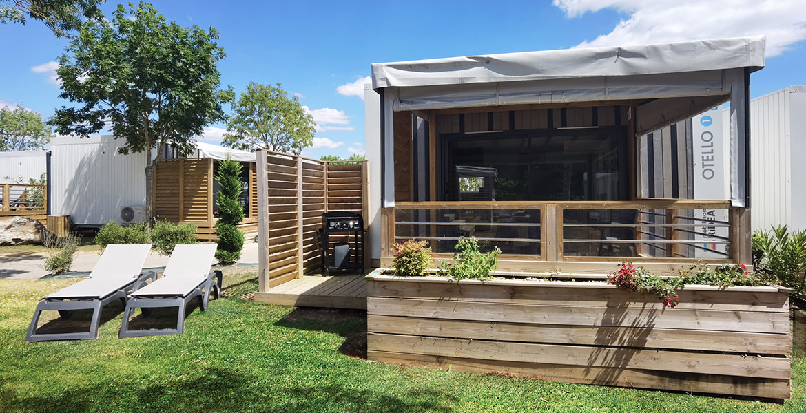 Terrasse extérieure d'un quartier Otello avec des chaises longues, un espace barbecue et une véranda en bois entourée de verdure sous un ciel bleu.