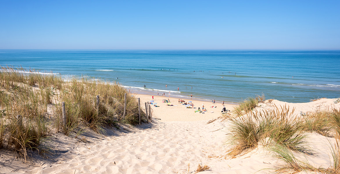 Chemin sablonneux entre des dunes menant à une plage animée avec des gens profitant du soleil et de la mer sous un ciel bleu clair.