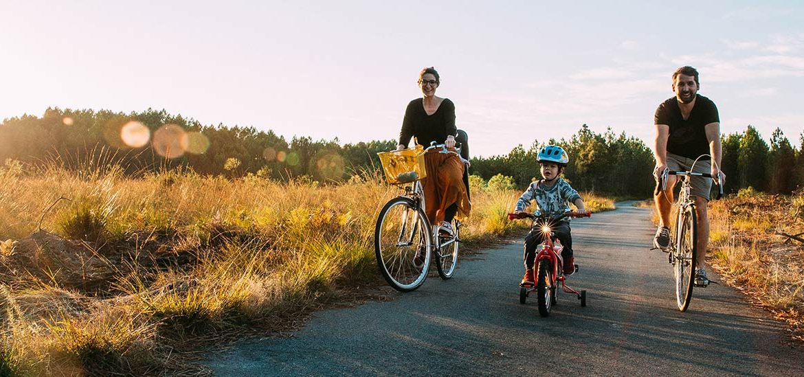 Famille faisant du vélo sur un chemin pavé à travers la campagne, avec des champs et des arbres en arrière-plan sous une lumière de coucher de soleil.