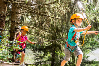Deux enfants portant des casques et des harnais de sécurité traversant un parcours d'accrobranche en forêt.